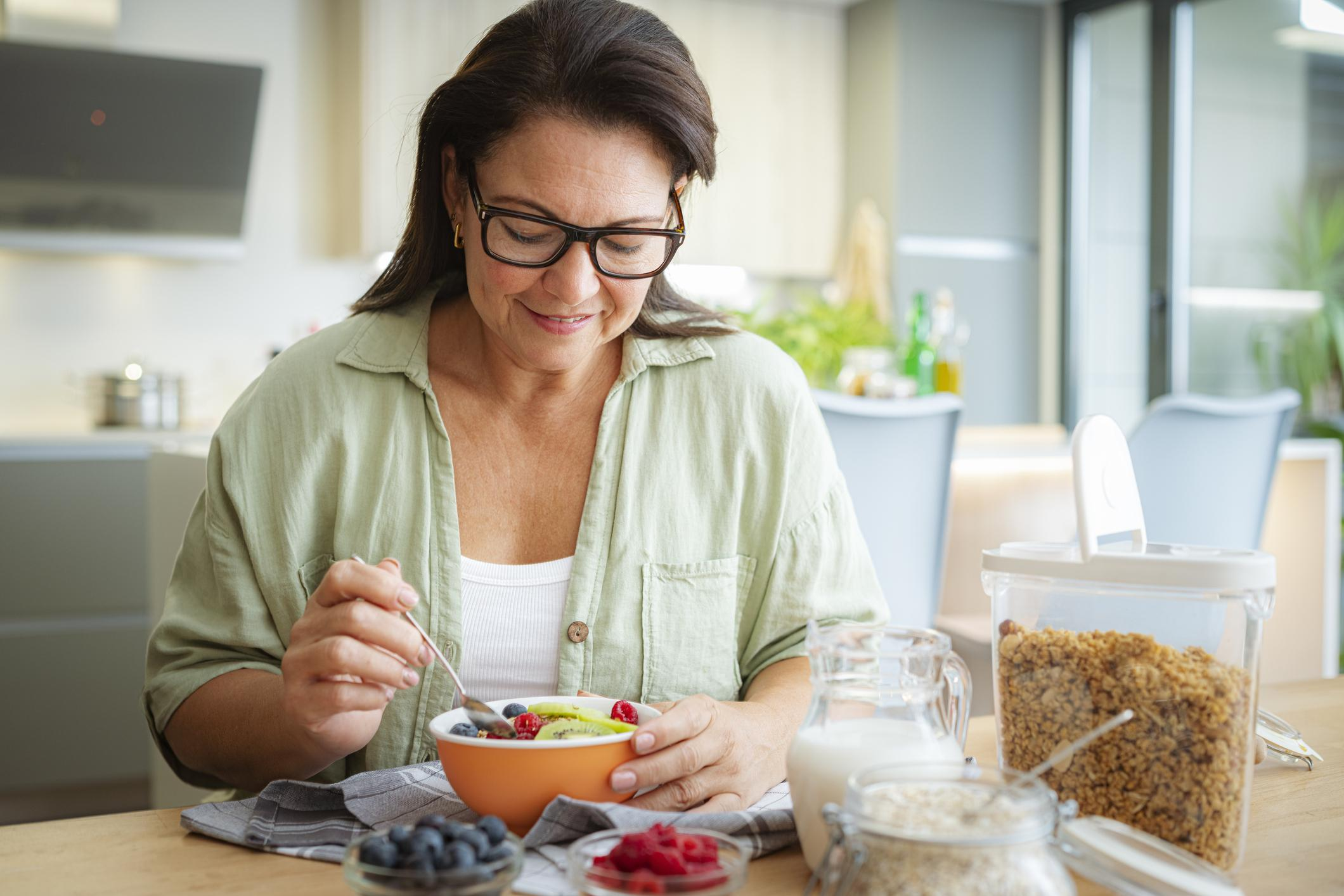 Happy mature woman eating fruits and granola sitting at kitchen table Happy mature woman eating fruits and granola sitting at kitchen table