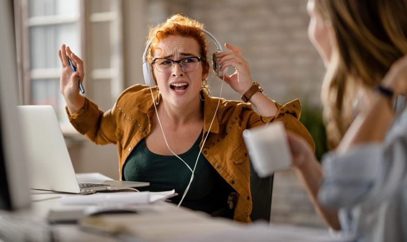 Creative business woman with headphones apologizing for not hearing her colleague talking to her while working in the office.