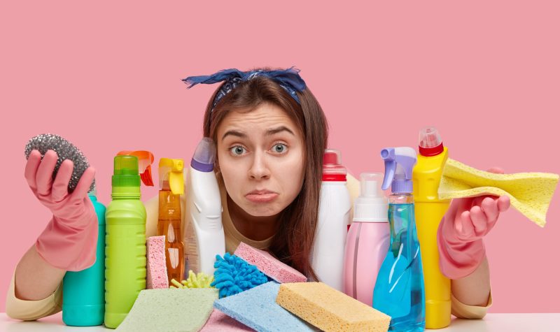 European clueless young housekeeper looks with apathy at camera, spreads palms, wears protective gloves, carries rag, models against pink background, surronded with multipurpose cleaning agents